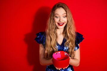 Young woman in blue sequin dress holding a round red gift box, stylish and cheerful with blonde...