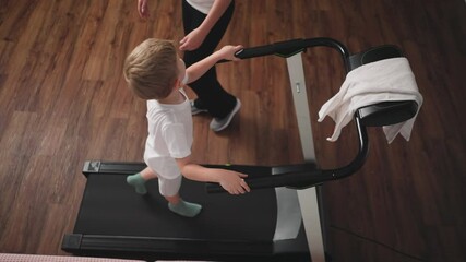 Overhead view of young boy walking on treadmill turning to look at aunt as she adjusts towel and prepares to operate machine in cozy home setting with wooden floor and soft lighting