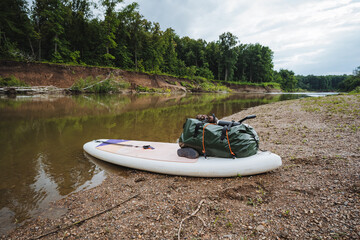 A paddle board that has a bag resting on it is positioned on the sandy shore of a river, ready for...