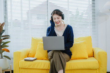 Young Asian woman sitting on yellow sofa listening to music happily relaxing in room