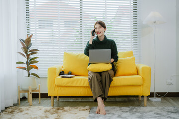 Freelance Asian woman holding mobile phone typing on laptop and working from home. Happy young girl sitting on sofa in living room.