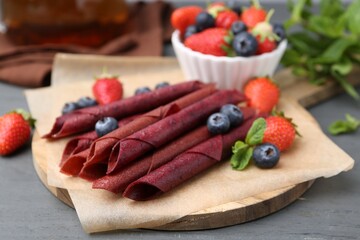 Tasty fruit leather rolls, strawberries, blueberries and mint on grey wooden table, closeup