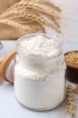 Wheat flour in glass jar, grains and spikes on white table, closeup