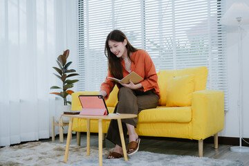 
Beautiful young woman sitting on a sofa and reading a book with a laptop, smartphone, iPad placed...