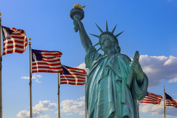 Fototapeta premium Statue of Liberty holds her torch high while American flags flutter in breeze, symbolizing freedom hope.