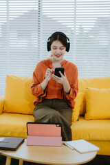 Fototapeta premium Young asian woman using smartphone and wearing headphone sitting on couch at home. Happy girl relaxing comfortable in the living room, spending lazy weekend, listening to music, watching movie.