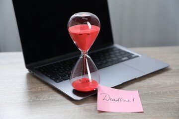 Sticky note with word Deadline, hourglass and laptop on wooden table indoors, closeup