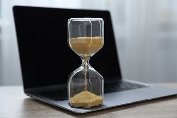 Deadline. Hourglass with flowing sand and laptop on wooden table indoors, closeup