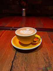 Latte art coffee served in a yellow cup and saucer, placed on a rustic wooden table in a cozy cafe setting