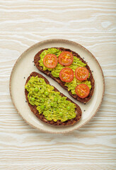 Two slices of whole rye bread are topped with mashed avocado, green peas, cherry tomatoes, and sesame seeds, a healthy and vibrant breakfast or snack option