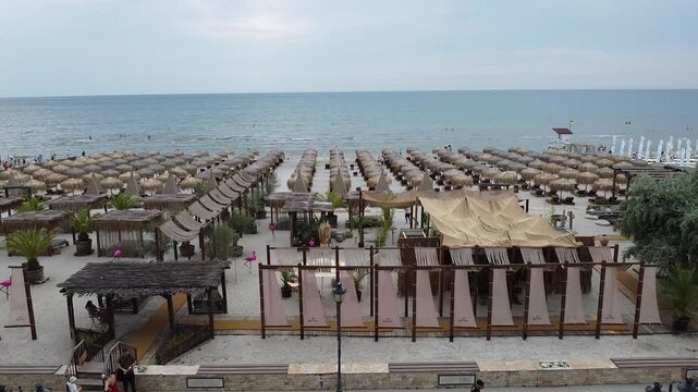 Drone view of beach scene with rows of thatched umbrellas, beachgoers and sunbeds, leading to ocean