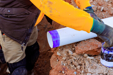 Worker is preparing section of PVC pipe with adhesive in trench for plumbing installation at...