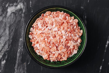 Pink Himalayan salt in green bowl. Natural coarse salt crystals closeup. Healthy seasoning mineral on black marble table. Raw organic ingredient for cooking, detox or skincare.