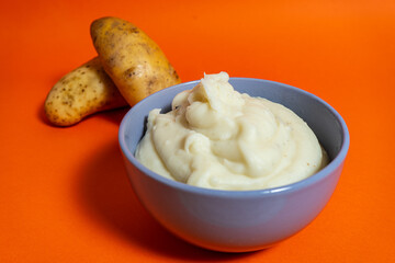 Creamy mashed potatoes with raw potatoes on vibrant orange background