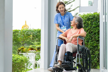 smiling Asian caregiver in scrubs enjoys a moment outdoors with senior woman in a wheelchair,...