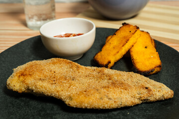 Breaded chicken breast with fried polenta and ketchup on dark plate
