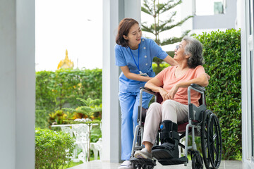 smiling Asian caregiver in scrubs enjoys a moment outdoors with senior woman in a wheelchair,...
