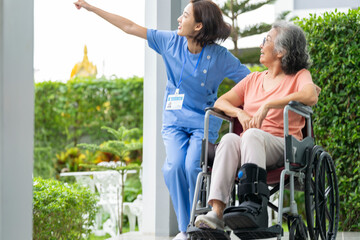 smiling Asian caregiver in scrubs enjoys a moment outdoors with senior woman in a wheelchair,...