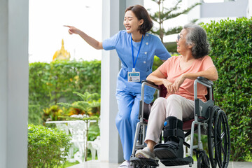 smiling Asian caregiver in scrubs enjoys a moment outdoors with senior woman in a wheelchair,...