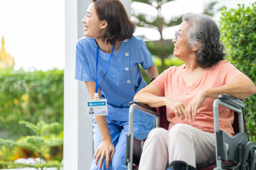 smiling Asian caregiver in scrubs enjoys a moment outdoors with senior woman in a wheelchair,...