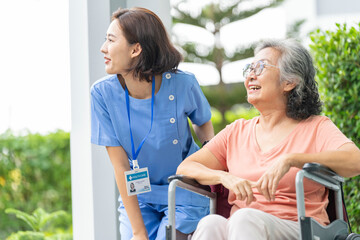 smiling Asian caregiver in scrubs enjoys a moment outdoors with senior woman in a wheelchair,...