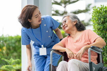 smiling Asian caregiver in scrubs enjoys a moment outdoors with senior woman in a wheelchair,...
