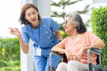 smiling Asian caregiver in scrubs enjoys a moment outdoors with senior woman in a wheelchair,...