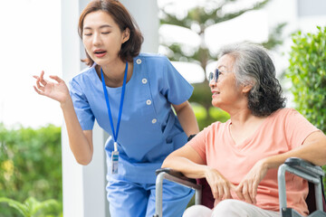 smiling Asian caregiver in scrubs enjoys a moment outdoors with senior woman in a wheelchair,...