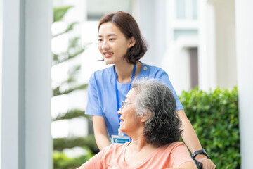 smiling Asian caregiver in scrubs enjoys a moment outdoors with senior woman in a wheelchair,...