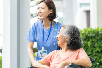 smiling Asian caregiver in scrubs enjoys a moment outdoors with senior woman in a wheelchair,...