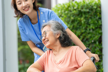 smiling Asian caregiver in scrubs enjoys a moment outdoors with senior woman in a wheelchair,...