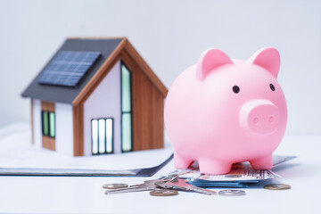 Pink piggy bank with house with solar panels on table with paper money, coins and house keys. Concept of saving for a house, real estate investment and eco-friendly housing.