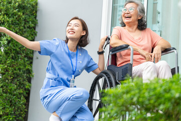 smiling Asian caregiver in scrubs enjoys a moment outdoors with senior woman in a wheelchair,...