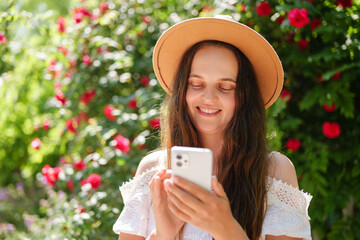 A joyful young woman in a brown hat and white dress smiles with eyes closed, engrossed in her...