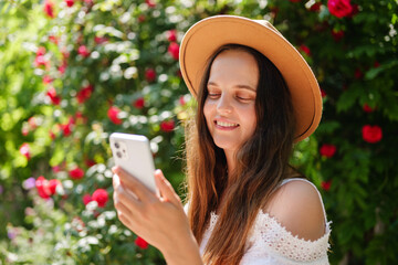 A young woman in a brown hat and white dress smiles, eyes closed in contentment, while scrolling on...