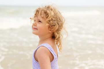 A joyful little girl with golden curly hair, wearing a purple swimsuit, smiles softly while looking...