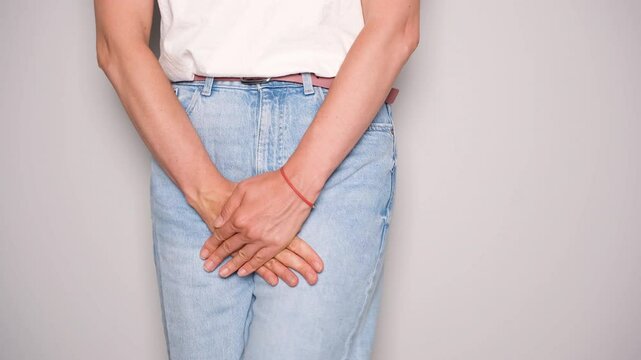 Close-up of a woman in jeans and white t-shirt standing with hands clasped in front of her lower abdomen, symbolizing the discomfort and urgency related to bladder leakage or urinary incontinence