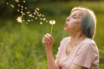 A senior woman smiling as she blows on a dandelion, captured in a peaceful outdoor setting with seeds drifting away.