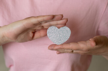 Close-up of female hands holding a shiny silver heart on a soft pink background, symbolizing love, kindness, and emotion
