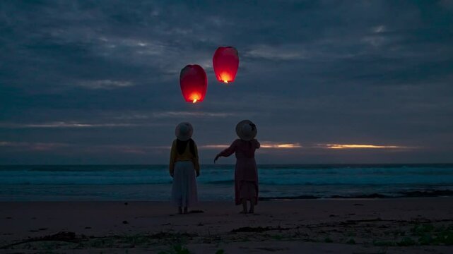 Two young women release lit paper lanterns at dusk on the beach. Two friends release paper lanterns into the sky