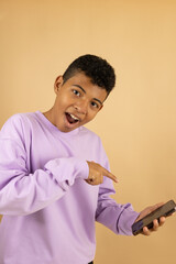Vertical image of a cheerful young Latino man wearing a lilac sweatshirt pointing at his cell phone on a light brown background with space to write advertising, promotion and offer concept