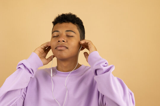 Brown skinned Latin American teenager wearing a lilac sweatshirt with his eyes closed feeling the rhythm, listening to music with wired headphones on a light brown background with copyspace