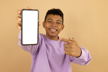 Latino teenager wearing a purple sweatshirt with a happy expression pointing at his cell phone with a blank screen for advertising space on a light brown background, Text area for advertising