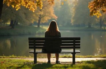 Fototapete Woman sits bench in park near lake. Solitude scene during autumn season. Girl enjoys peaceful moment in nature, quiet contemplation near water. Concept of alone time. Trees with yellow leaves, water © miss irine
