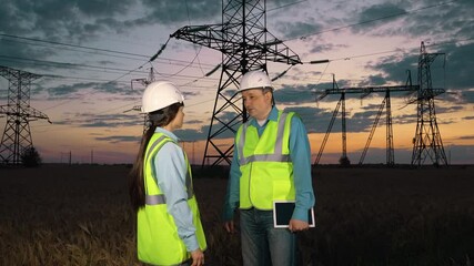Skilled engineer with tablet shakes hand of woman assistant at energy distribution substation at countryside. Electricians with tablet perform agreement at power generation plant in sunset field