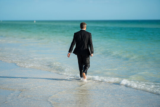 Leisure weekend and remote freelance work. Crazy comic business man in suit rest on sea bech. Rear view of back business man in suit in sea water at beach. Remote summer business. Summer holiday.