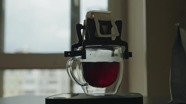 Close-up of coffee drip bag brewing fresh cup of coffee in glass mug placed on electronic kitchen scales, adding touch of precision to morning routine in modern kitchen setting by window, slow motion.