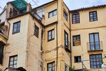 Historic residential building with cracked yellow stucco walls, wooden window frames, wrought iron balconies, and traditional roof tiles; architecture, facade;