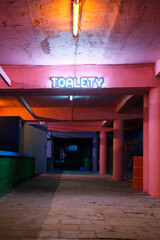 Neon-lit underground passage with a glowing “TOALETY” sign above, indicating restrooms. Pink and orange hues create a retro, moody atmosphere.