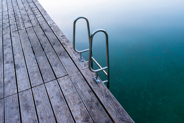 Top-down view of a wooden deck with a stainless steel ladder leading into transparent turquoise lake water. Peaceful summer setting ideal for design, travel, minimalism, or wellness themes.